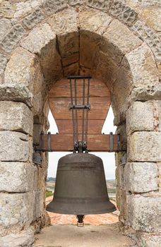 Aged metal bell in arched hole of stone brick wall of ancient castle near sea in Spain in sunny day