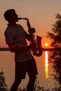 A young guy plays the saxophone in the evening at sunset.
