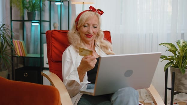 Senior old grandmother woman using credit bank card and laptop computer while transferring money purchases online shopping, order food delivery at home. Elderly lady pensioner sitting on chair at home