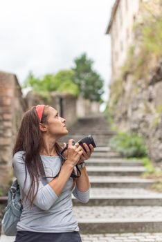 Side view of thoughtful Hispanic female tourist with long dark hair and backpack walking on aged stairway and taking photo of ancient stone building, in Spain while looking away