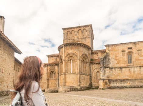 Side view of unrecognizable female tourist with long dark hair and backpack, admiring view of aged historic stone Colegiata de Santa juliana Santillana del Mar in Cantabria in Spain.
