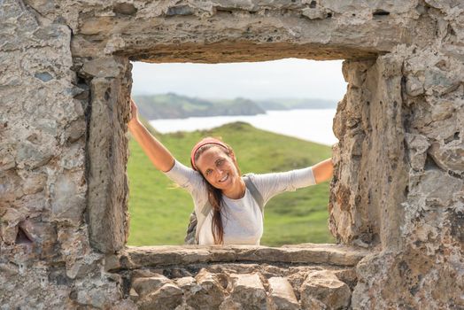 Smiling Hispanic woman standing on green hill near sea and leaning on aged stone building while looking at camera through stone hole in Spain