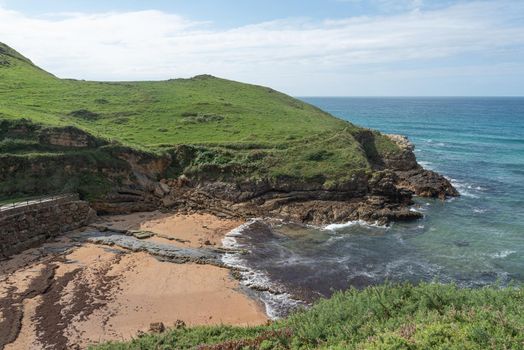 Amazing scenery of rocky cliff covered with green grass located near calm turquoise ocean water in sunny day in Cantabria in Spain