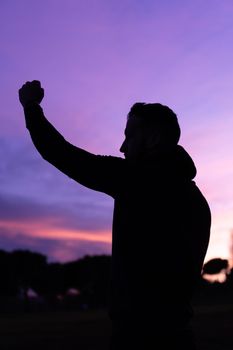 Side view silhouette of anonymous male raising fist up while celebrating success in cloudy evening at sundown