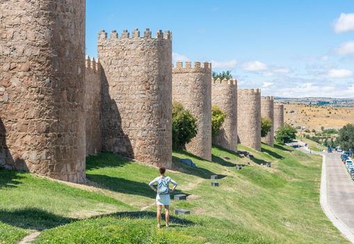 Back view of unrecognizable traveling female standing near aged Avila Walls with stone towers on sunny day in Spain