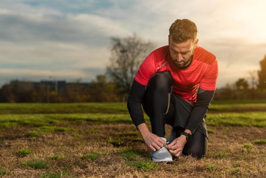 Serious bearded male jogger in sportswear kneeling on grassy ground and tying shoelaces on sneakers during running workout