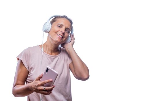 Front view of a cheerful adult woman listening music with her headphones and smartphone looking at camera on a white background with copy space.