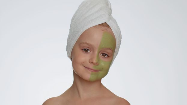 Beautiful young smiling child girl in towel on head applying cleansing moisturizing green mask on face. Teenager kid face skin care treatment, natural cosmetics. Female portrait. Perfect fresh clean