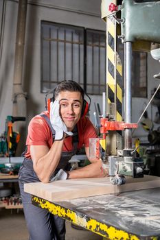 Smiling male woodworker in protective headphones and gloves leaning on band saw while working in carpentry workshop and looking at camera