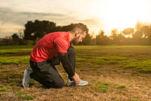 Full body side view of bearded male runner in sportswear tying laces on sneakers during jogging workout in park at sundown