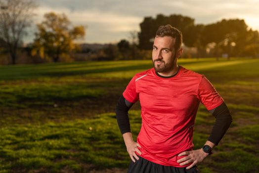 Bearded male athlete in sportswear standing with hands on waist in park during fitness training and looking at camera