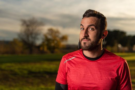 Bearded male athlete in sportswear raising eyebrows while looking at camera and listening to music in earphones during training