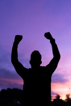 Silhouette of anonymous male raising fists and celebrating successful achievement in nature at sunset under bright cloudy sky