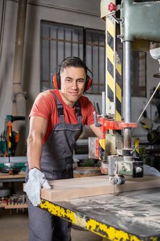 Positive male woodworker in uniform cutting wooden board on band saw and looking at camera with smile