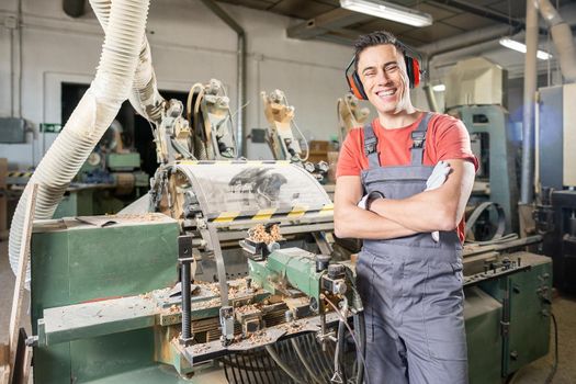 Positive male woodworker in protective headphones and uniform standing near electric wood turning machine in carpentry workshop and smiling at camera