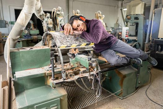 Full body of weary male carpenter in protective headphones having nap on wood turning machine after tiring workday in joinery workshop