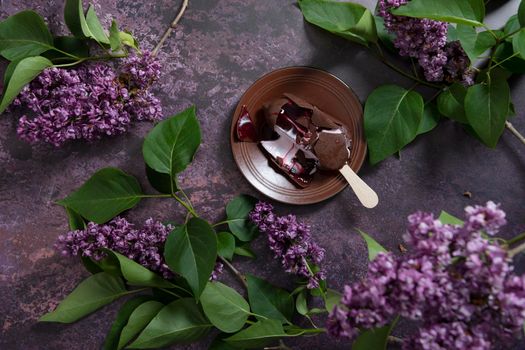 melted raspberry ice cream in chocolate on a wooden stick on a dark burgundy background with lilac flowers, a sweet dessert on a hot spring day, flat lay food. High quality photo