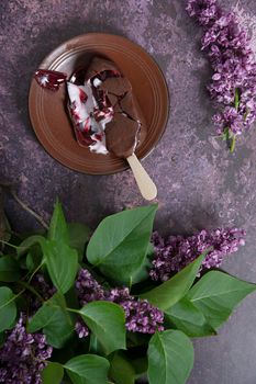 melted raspberry ice cream in chocolate on a wooden stick on a dark burgundy background with lilac flowers, a sweet dessert on a hot spring day, flat lay food. High quality photo