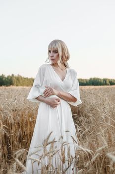 A blonde woman in a long white dress walks in a wheat field. The concept of a wedding and walking in nature.