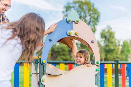 Front view of a happy little blond boy playing inside a wooden play set in the playground with his two adoptive parents.