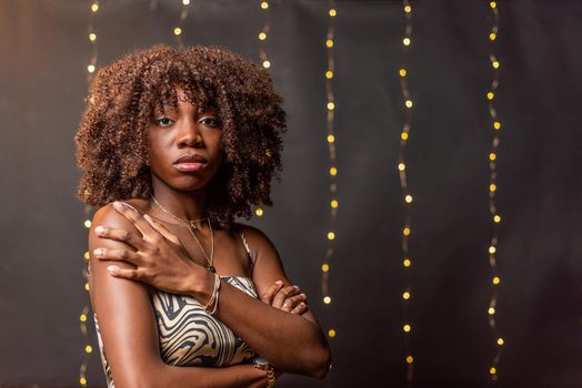 Unsmiling African American young woman with curly hair looking at camera posing with hand on shoulder on a blurred light background.