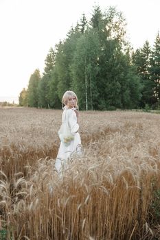 A blonde woman in a long white dress walks in a wheat field. The concept of a wedding and walking in nature.