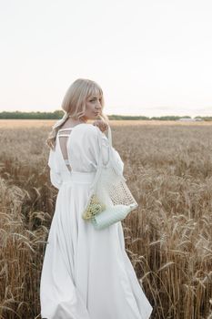 A blonde woman in a long white dress walks in a wheat field. The concept of a wedding and walking in nature.