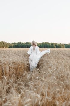 A blonde woman in a long white dress walks in a wheat field. The concept of a wedding and walking in nature.