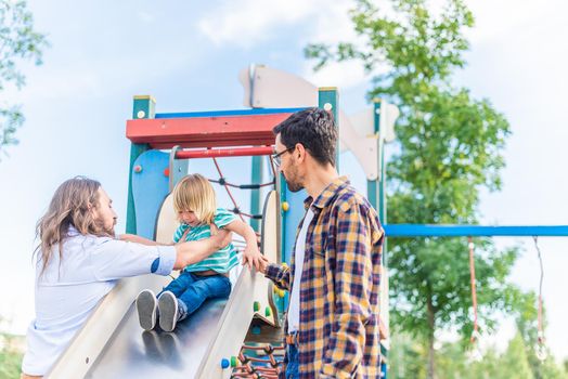 Front view of a gay male couple with their adopted son teaching him how to play on the slide at a playground.