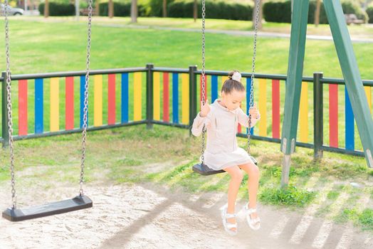 Sad little girl sitting alone on a swing looking down in a playground. Concept of loneliness, girl with no friends.