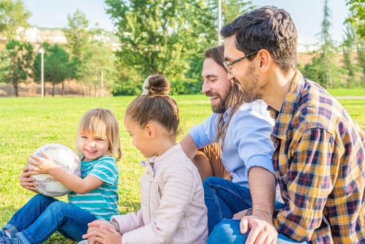 Side view of a gay male couple with their two children sitting on the grass in a park having a good time as a family.