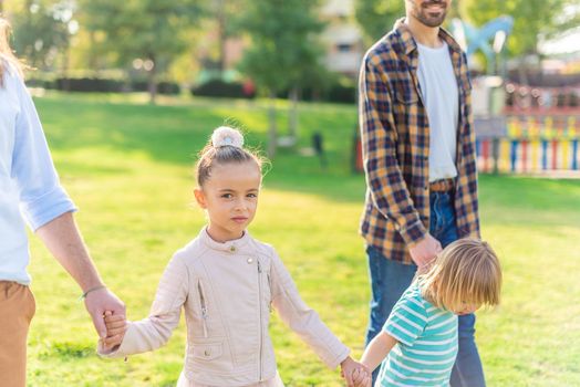 Portrait of a little girl looking at the camera holding hands with her little brother and her two adoptive dads in the park.