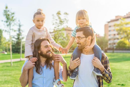 Front view of a young gay male couple holding on their shoulders their children holding hands and looking at camera in the park