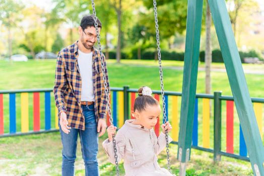 A young man playing with his adopted daughter on the swing in a playground in a sunny day.