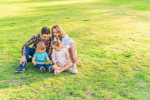 Full length view of a young gay male couple with their adopted children sitting on grass in the park reading a children's book