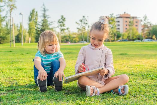 Front view of a little girl and little brother sitting on the grass in a park having fun drawing on a small children's board.