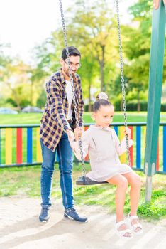 Vertical image of a young man playing with his adopted daughter on the swing in a playground in a sunny day.