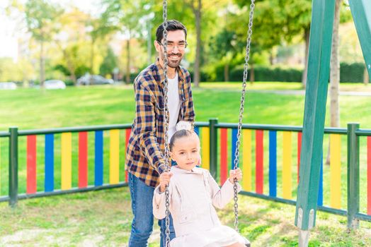 A young man playing with his adopted daughter on the swing looking at camera in a playground in a sunny day.