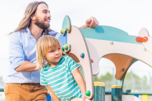 Front view of a little blond boy playing on a wooden game on the playground with his young father looking both away