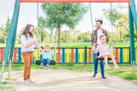 Front view of a happy young gay couple playing with their children on the swing at the playground in a sunny day.