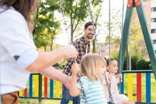 Side view of a happy young gay couple looking at each other playing with their children on the swing.