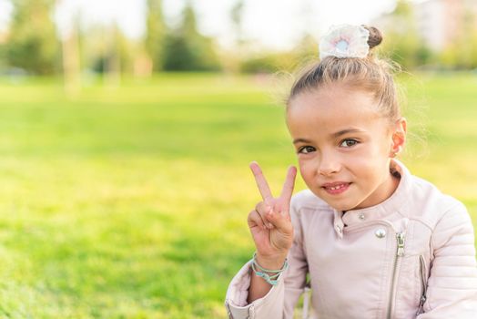 Portrait of a little girl sitting on grass in a park looking at camera and showing peace or victory sign with copy space