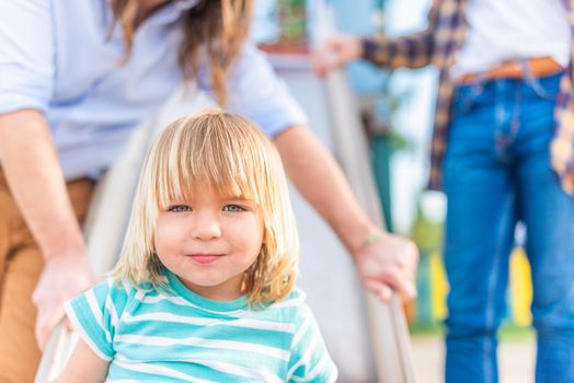 Portrait of a happy blond boy looking at the camera sitting on a playground slide with his two unrecognizable dads behind him.