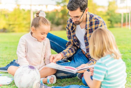 Front view of a young single father reading a book for children to his children sitting on the grass in a park. Concept of single parent family.