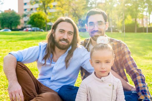 Portrait of gay male couple with their daughter sitting on grass of a park.
