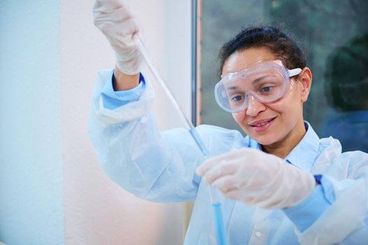 Experienced multi-ethnic woman chemist analyst, scientist wearing safety goggles and white lab coad holds a laboratory pipette in her gloved hand and titrates chemical substance into a test tube