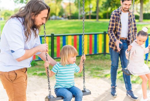 A young gay couple playing with their children on the swing at the playground in a sunny day.