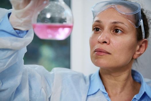 Multi-ethnic middle-aged woman scientist chemist in white lab coat and goggles, works with liquids in labware, analyzes flask with pink fluid substance in a chemistry research laboratory