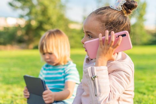 Side view of a little girl talking on a smart phone sitting on the grass with her little brother holding a digital tablet