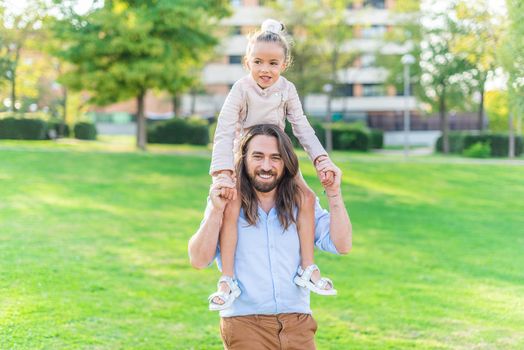 Front view of a happy young father holding her little daughter on his shoulder in park